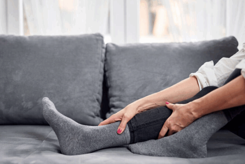 A person sitting on a couch, wearing grey socks and holding their lower leg with both hands as if massaging or soothing discomfort.
