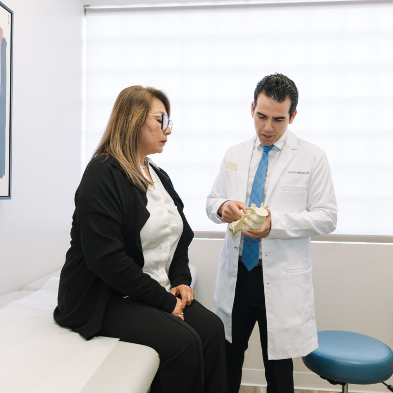 “Doctor in a white coat explaining a spine model to a seated patient during a consultation in a modern medical exam room.”